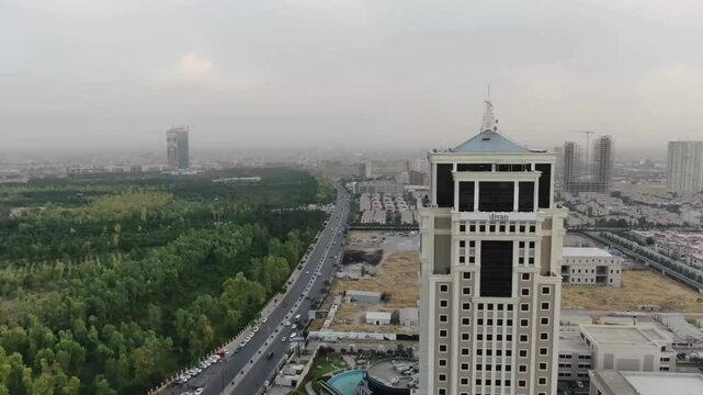 Aerial drone view of the Divan Tower in a developing urban area with highway, green parkland, and construction sites under hazy daylight