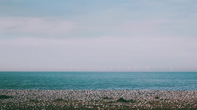 Calm pebble beach with turquoise sea and distant wind turbines on the horizon under soft overcast sky