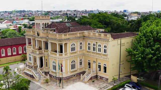 Low frontal aerial drone approach to the ornate yellow facade of Palacio Rio Negro, former governor's palace of Manaus, arched windows, columns, flags and the red annex beside it, overcast, Brazil.