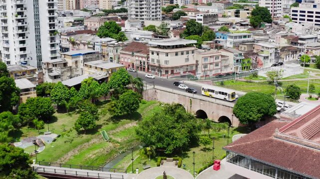 Aerial drone tracks past an arched stone bridge with cars and a bus crossing the igarape stream to the red cycleway, walkways and lawns of Parque Jefferson Peres, sunny day, Manaus, Amazonas, Brazil.