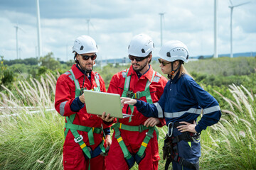 Fototapeta premium A team of engineers in safety gear gathers outdoors to review data on a laptop computer. They discuss a project plan while working near a wind farm field under an open sky.