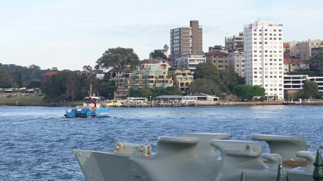 Blue environmental services workboat travels across Sydney Harbour with waterfront apartment buildings and residences behind. Sydney, New South Wales, Australia.
