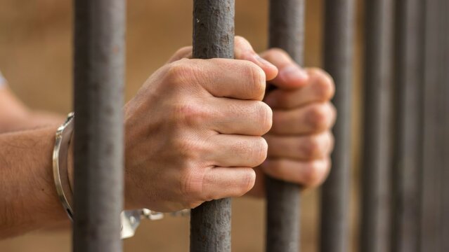 Close-up of handcuffed hands gripping vertical metal prison bars, blurred background, human skin texture, metallic texture of shackles and bars
