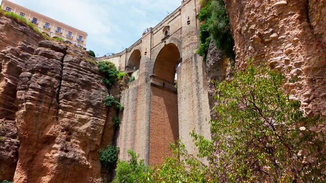 Upward and downward pan beneath Ronda&rsquo;s famous Puente Nuevo, revealing the dramatic El Tajo gorge, rocky cliffs and lush greenery below.