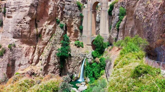 Upward pan beneath Puente Nuevo revealing the waterfall, deep El Tajo gorge, dramatic cliffs and surrounding greenery in daylight.