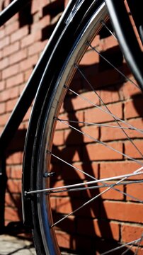 Black bicycle stands motionless beside sunlit red brick wall as view moves progressively closer to front wheel, narrowing from full frame and saddle to rim, spokes, tire, and hard-edged shadow