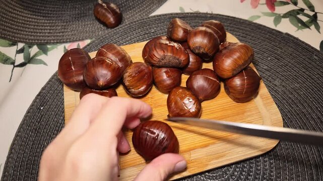 Hands carefully cutting a chestnut with a knife on a wooden board, surrounded by a pile of glossy fresh chestnuts