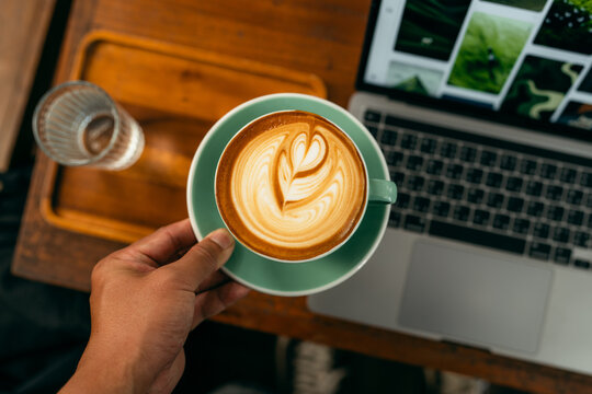 POV of freelancer holding coffee cup while browsing social media and photos on laptop in a cafe.