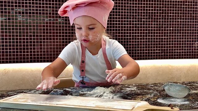 Horizontal 16:9 video. Medium close-up: a little girl wearing a white T‑shirt and a pink chef hat with apron straps works at a dark stone countertop