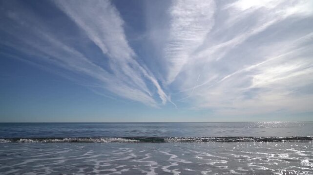 Gentle ocean waves rolling onto a sandy beach under a vast blue sky with streaky cirrus clouds