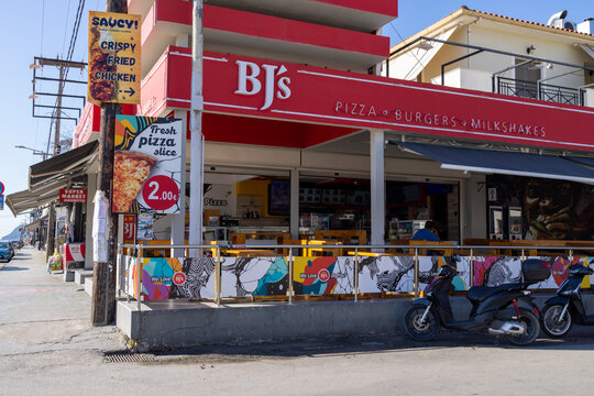 Laganas, Zakynthos Greece, 1st September 2026: BJ's pizza burgers and milkshakes restaurant frontage with colourful signs outdoor seating scooters and food advertising on a busy Greek resort street