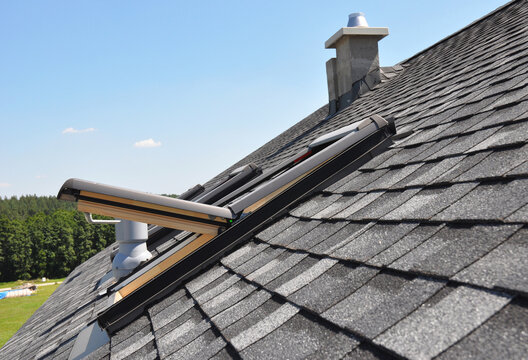 Skylight on asphalt shingle house roof with chimney and ventilation pipe.