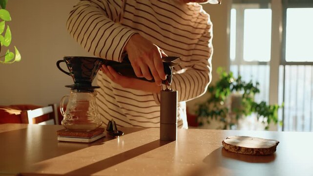 Man grinding fresh roasted coffee beans with a manual grinder in a sunlit kitchen for his morning pour over coffee ritual, preparing a delicious and aromatic beverage