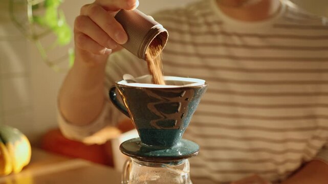 Barista pouring freshly ground coffee from a manual grinder into a dripper with a paper filter, preparing a morning beverage with the pour over method in a warm, sunlit kitchen