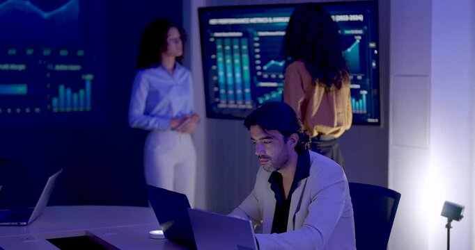 Adult male professional types and smiles at laptop while two female colleagues discuss performance dashboard behind him, combining independent computer work with collaborative business data analysis.