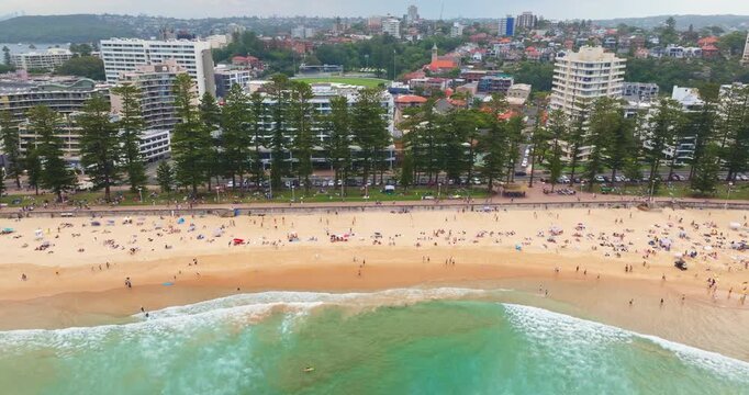Aerial view of Manly Beach in Sydney, Australia, turquoise ocean waves breaking on the sandy shore, people relaxing, swimming, and surfing n summer weekend. Coastal buildings houses line the promenade