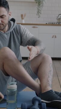 Young man in sportswear using smartphone then training with dumbbells sitting on yoga mat in apartment. Domestic workout and modern technology concept.