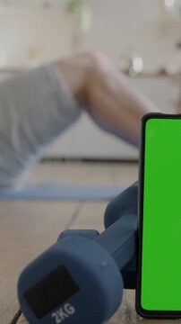 Close-up of smartphone with chroma key green screen near dumbbells and sportsman doing crunches at home in background. Sports and mobile application concept.