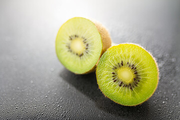 Two sliced fresh kiwi halves with brown fuzzy skin and green flesh, placed on a dark surface with water droplets. Close-up view of ripe fruit. © tai