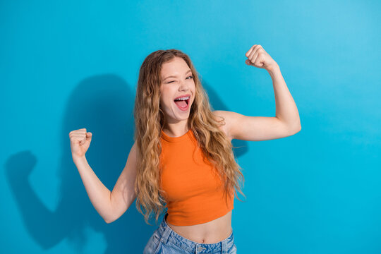 Young glamorous girl with orange top flexing arms against blue background smiling and feeling confident