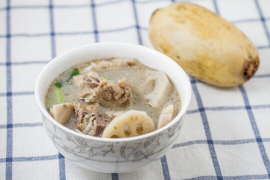 A bowl of simmered lotus root and pork ribs soup, with a whole raw lotus root beside it on a checkered tablecloth.
