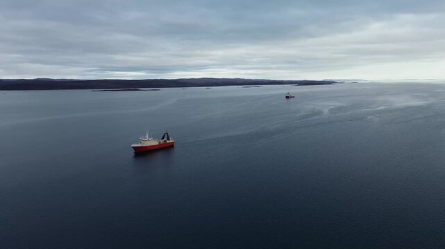 Mid altitude aerial drone tracking shot of red vessel Mersey Venture sailing parallel to distant Arctic island shores with Captain Molly Cool anchored nearby