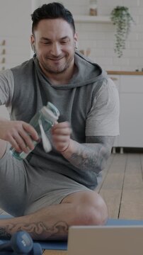 Young man in sportswear talking to online fitness trainer during video call discussing workout and drinking water indoors at home. Communication and virtual meeting concept.