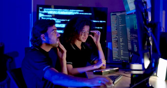 Adult male and female technology professionals review source code on monitors, pause to discuss findings and continue computer work during cybersecurity analysis in modern digital workplace.