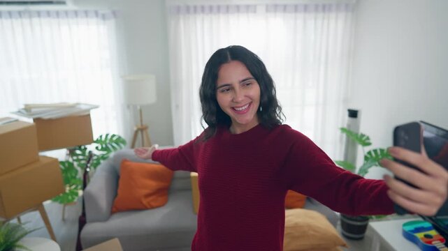 Young Hispanic woman using phone video call while moving into a new home. 