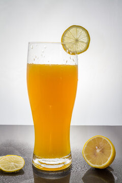 A tall transparent glass filled with bright orange juice, garnished with a lemon slice, with additional lemon slices on the dark surface below.
