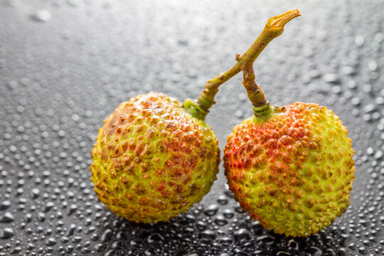 Two fresh lychees with bumpy red-green shells rest on a water-dotted dark surface, showing their stems and glossy texture.