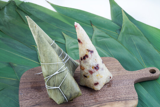 Two triangular zongzi, one wrapped in bamboo leaves, one unwrapped, placed on a wooden board with bamboo leaves in the background.