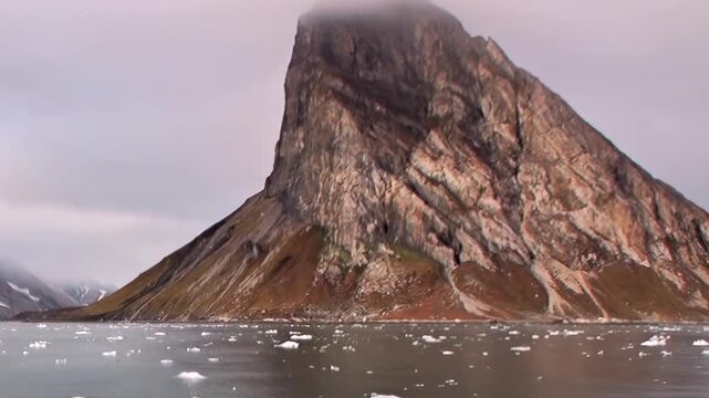 A steep rocky mountain peak rises above cold coastal water. The image retains wide framing and ice-strewn water as secondary detail, while light and texture shape the wider scene.