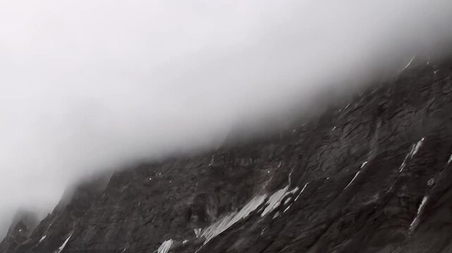 A steep mountain slope rises into fog above cold coastal water. Visible secondary elements include close framing and snow patches, adding depth and context to the overall composition.