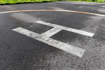 Obraz premium Helipad landing marking painted on asphalt tarmac showing a weathered white H and faded yellow circle, conveying aviation, emergency readiness, texture and urban outdoor infrastructure