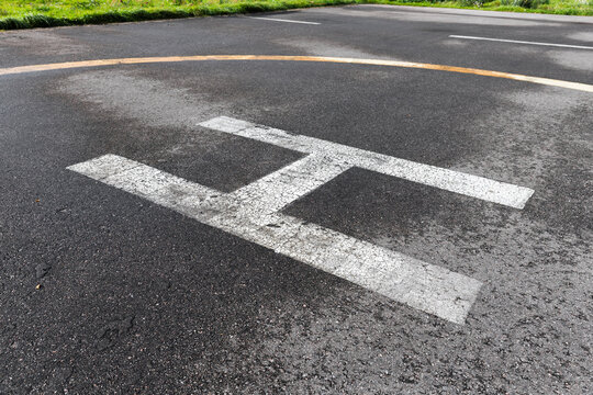 Helipad landing marking painted on asphalt tarmac showing a weathered white H and faded yellow circle, conveying aviation, emergency readiness, texture and urban outdoor infrastructure