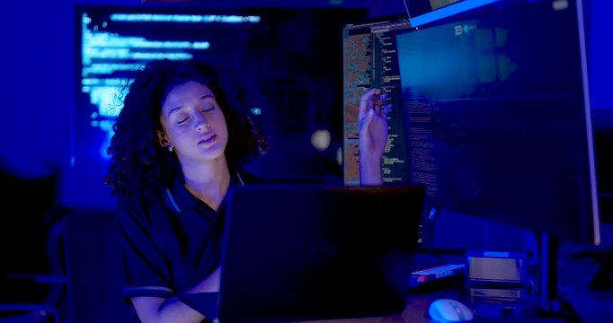 Adult female technology professional works on laptop, speaks and gestures while reviewing source code across monitors during cybersecurity analysis and system monitoring in digital operations office.