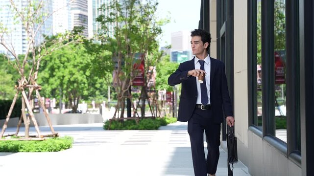 Businessman walking alone to work along a glass office facade, glancing at his watch as he keeps pace with the morning rush on a tree-lined walkway in central Bangkok