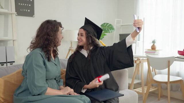 Young Latino daughter wearing graduation gowns doing video call with mom. 