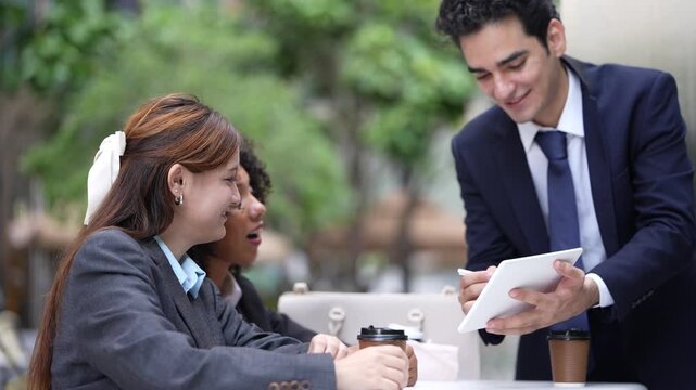 Three colleagues meet over a tablet at a shaded outdoor table, the man presenting while both businesswomen lean in, a compact alfresco project review in the city centre