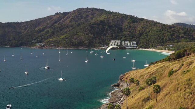 Windmill Viewpoint in Phuket presents active outdoor sports. Paraglider soaring above bay with many yachts anchored in clear turquoise waters and a lush green hillside framing the tropical coastline