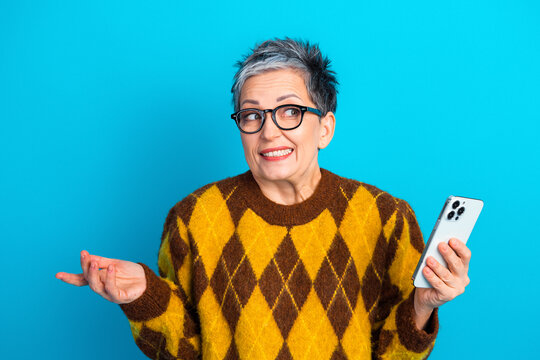 Senior woman with smartphone looking confused and amused in yellow argyle sweater and glasses against bright blue background
