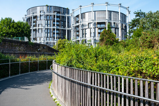 Curving pedestrian walkway with metal railing and greenery leads toward modern residential buildings, combining landscape, circulation and urban architecture.