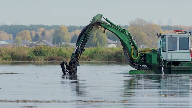 Dredger floating excavator digs river bottom with scoop to mine silt. Powerful drainage machine works with underwater deposits at riverside