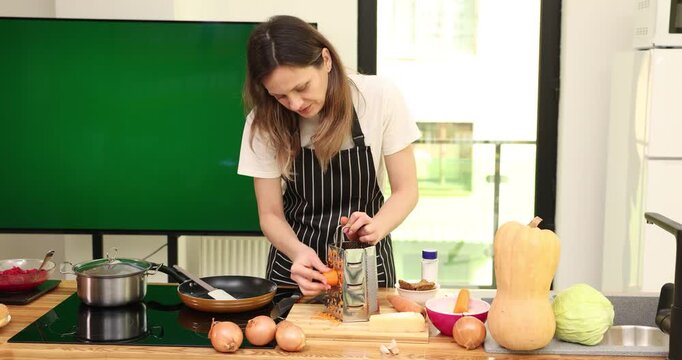 Home cook in striped apron works near vegetables on kitchen counter. Woman rubs fresh carrot across metal grater preparing ingredient for family meal