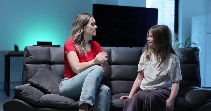 Mother scolds naughty little girl sitting nearby on black sofa at home. Woman talks to playful preteen daughter to discuss behavior in room