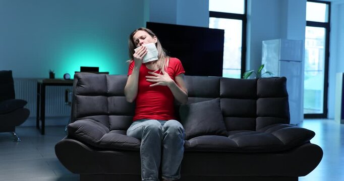 Pensive woman coughs covering mouth with napkin in living room. Blonde female with bronchitis symptoms sits on sofa at home. Flu disease slow motion