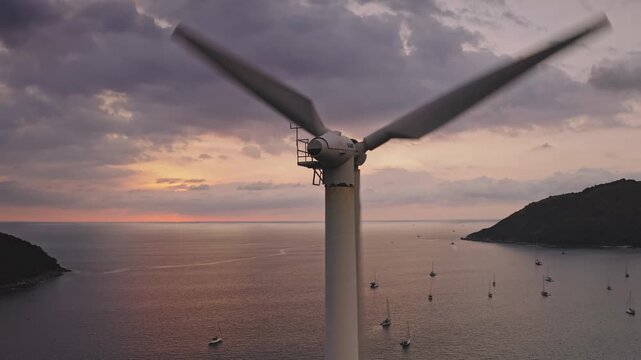 Close-up drone flight shot of wind turbine spinning its blades, generating renewable clean energy over the Andaman Sea at sunset, with many sailboats anchored around a bay near Phuket Island, Thailand