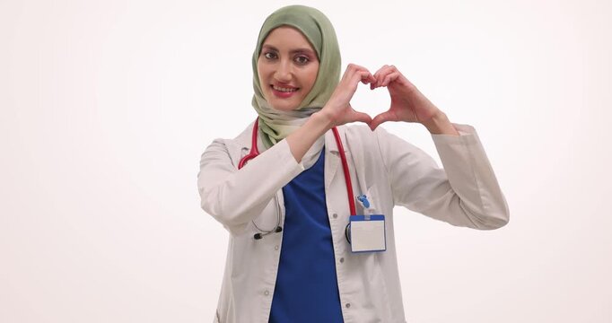 Cheerful woman cardiologist in hijab and white medical coat in clinic. Muslim heart specialist forms heart shape with hands against white background