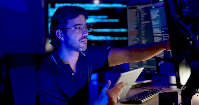 Adult Asian male cybersecurity analyst holds tablet and reaches toward monitor while reviewing source code, combining touchscreen interaction with network security monitoring and secure data analysis.